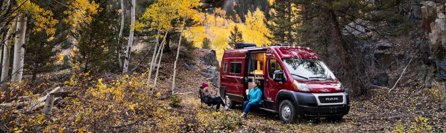 A couple of people sit outside of their Winnebago Solis RV while sipping drinks in the midst of Autumn.